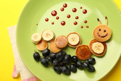 Creative serving for kids. Plate with cute caterpillar made of pancakes, grapes and banana on yellow background, top view