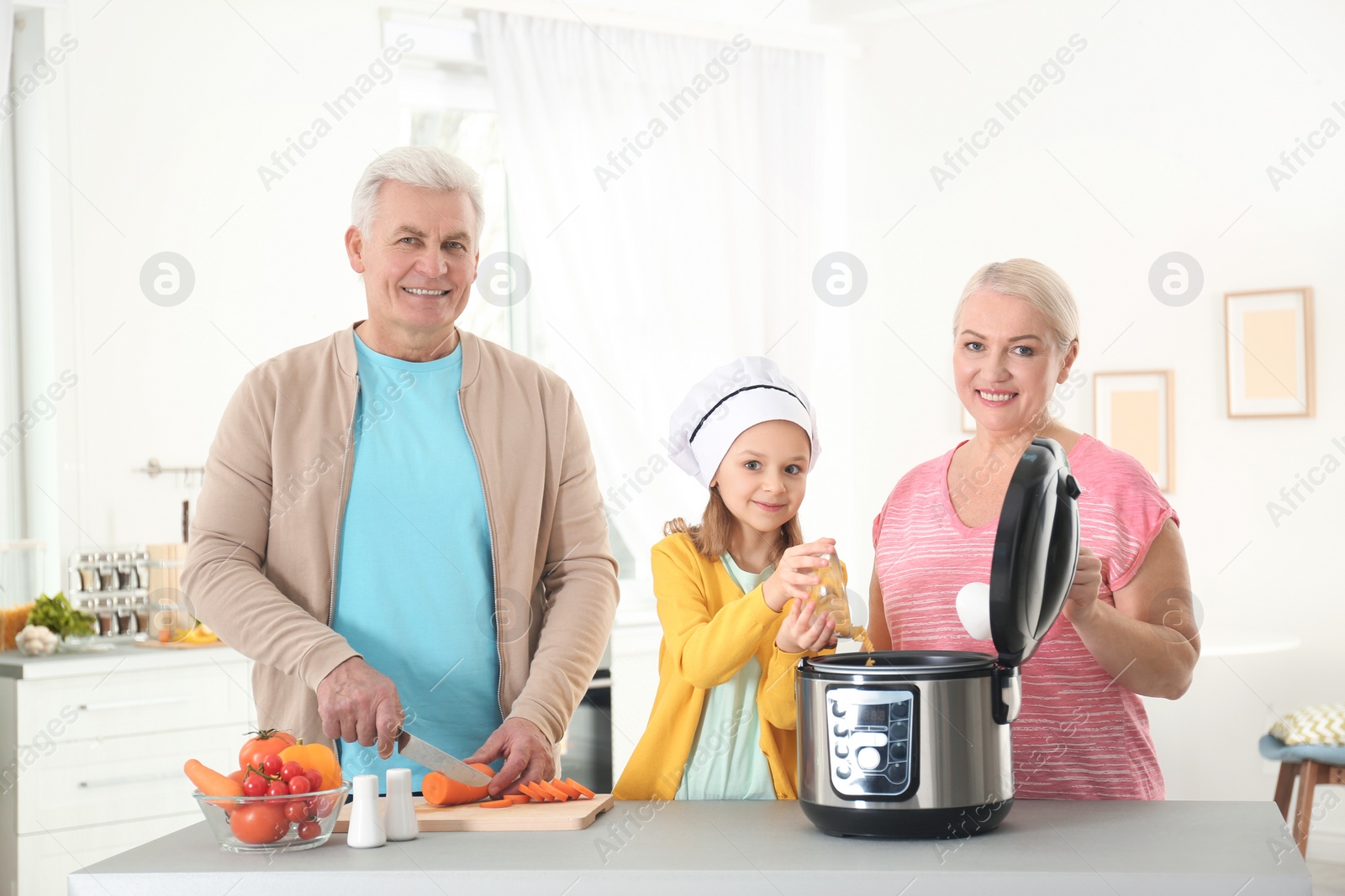 Photo of Mature couple and their granddaughter preparing food with modern multi cooker in kitchen