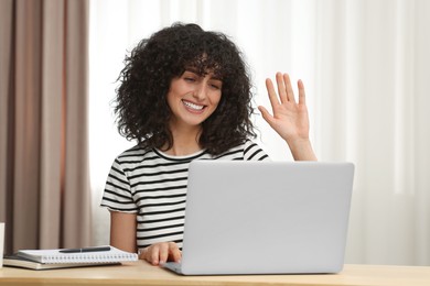 Photo of Happy woman waving hello during video call at table indoors