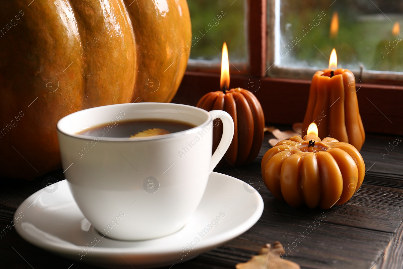 Photo of Cup of hot drink and pumpkin shaped candles on wooden table near window, closeup. Cozy autumn atmosphere