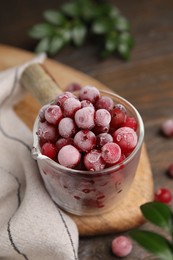 Photo of Frozen red cranberries in glass pot and green leaves on wooden table, closeup