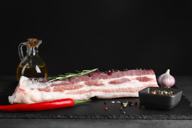 Photo of Piece of raw pork belly, rosemary, chili pepper and spices on grey table