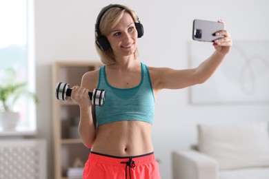 Photo of Smiling sports blogger holding dumbbell while streaming online fitness lesson with smartphone at home