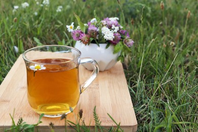 Cup of aromatic herbal tea and ceramic mortar with different wildflowers on green grass outdoors. Space for text