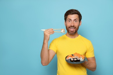 Photo of Emotional man holding tasty sushi roll with chopsticks on light blue background. Space for text