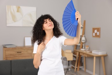 Photo of Young woman waving blue hand fan to cool herself at home