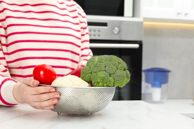 Photo of Woman holding colander with fresh vegetables at white marble table in kitchen, closeup. Space for text