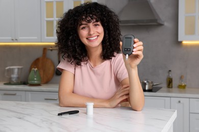 Photo of Diabetes. Happy woman holding digital glucometer at white marble table in kitchen