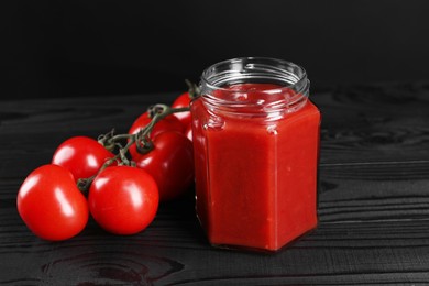 Photo of Delicious ketchup in jar and tomatoes on black wooden table, closeup. Tomato sauce