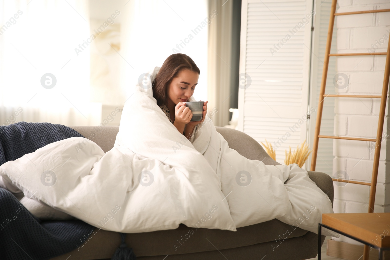 Photo of Beautiful young woman wrapped with soft blanket holding cup of hot drink on sofa at home