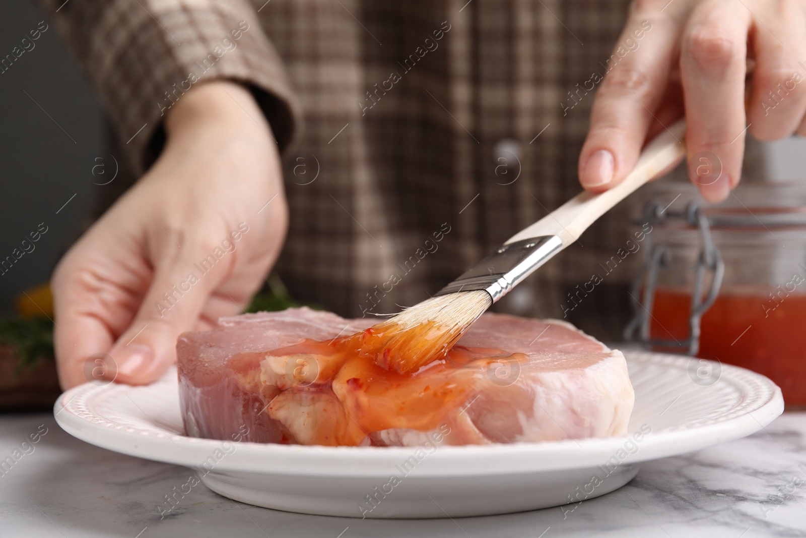 Photo of Woman spreading marinade onto raw meat with basting brush at white marble table, closeup