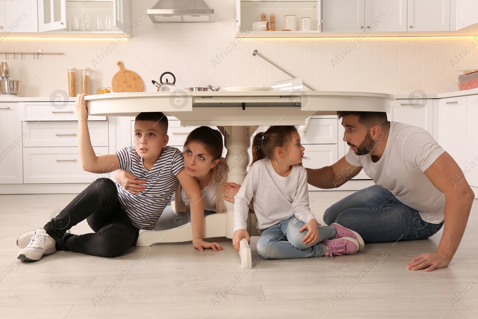 Photo of Scared parents with their children hiding under table in kitchen during earthquake