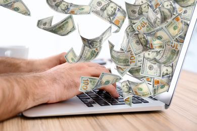 Image of Man with modern laptop and flying dollar banknotes at table, closeup. People make money online