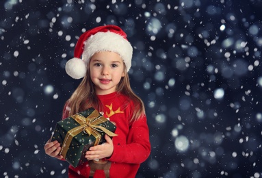 Image of Cute child in Santa hat with Christmas gift under snowfall on dark background