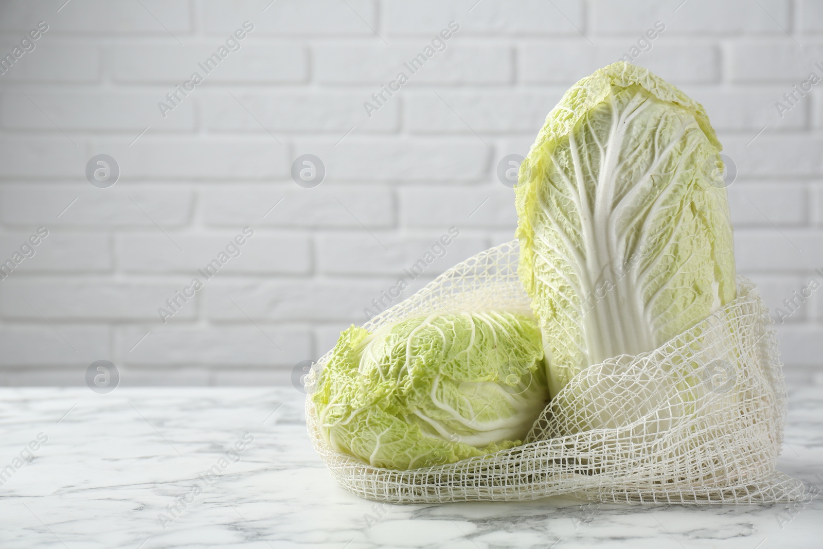 Photo of Fresh Chinese cabbages in string bag on white marble table near brick wall. Space for text