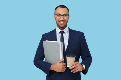 Photo of Happy young intern holding notebooks, takeaway cup with hot drink and muffin on light blue background