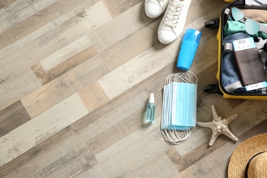 Flat lay composition with suitcase, protective masks, antiseptic spray and personal items on wooden floor, space for text. Travelling during coronavirus pandemic