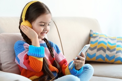 Photo of Cute child with headphones and mobile phone on sofa indoors
