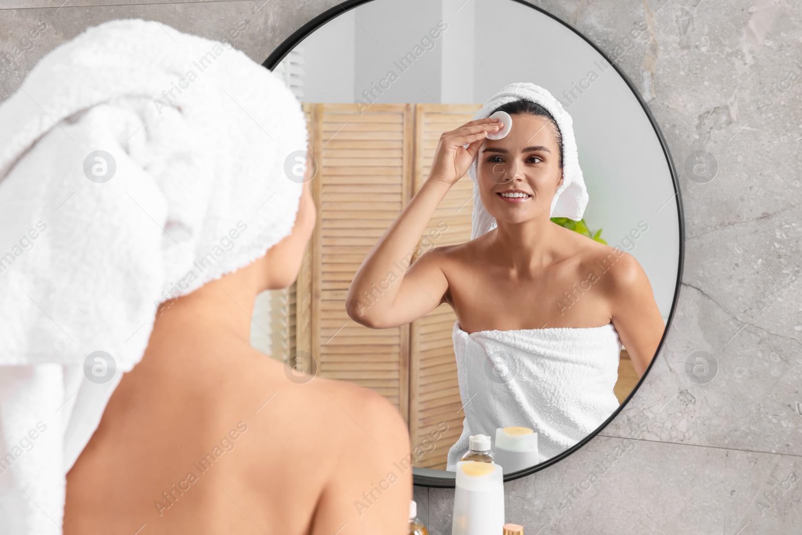 Photo of Young woman cleaning her face with cotton pad near mirror in bathroom