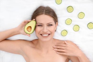 Photo of Beautiful woman with cucumber slices and avocado on white fabric, above view. Organic face mask
