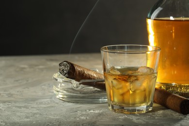 Photo of Bottle, glass of whiskey with ice cubes and cigars on grey table, closeup. Space for text