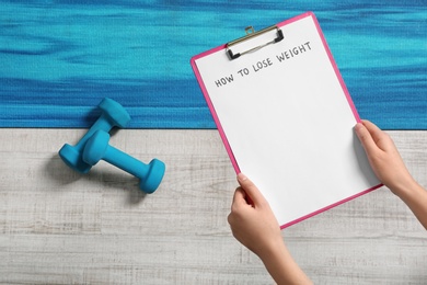 Photo of Woman holding clipboard with words HOW TO LOSE WEIGHT over sport equipment, top view