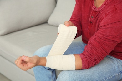 Photo of Young man applying bandage on injured arm at home, closeup. First aid