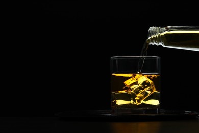 Pouring whiskey from bottle into glass with ice cubes at table against black background, space for text