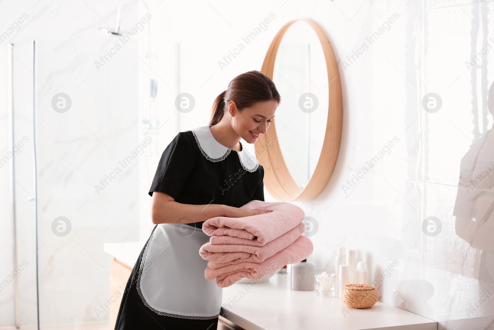 Photo of Young chambermaid putting stack of fresh towels on countertop in bathroom