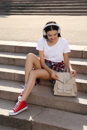 Photo of Beautiful young woman with stylish beige backpack and headphones on stairs outdoors