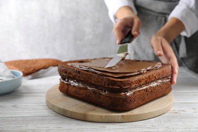 Photo of Woman smearing sponge cake with chocolate cream at white wooden table, closeup