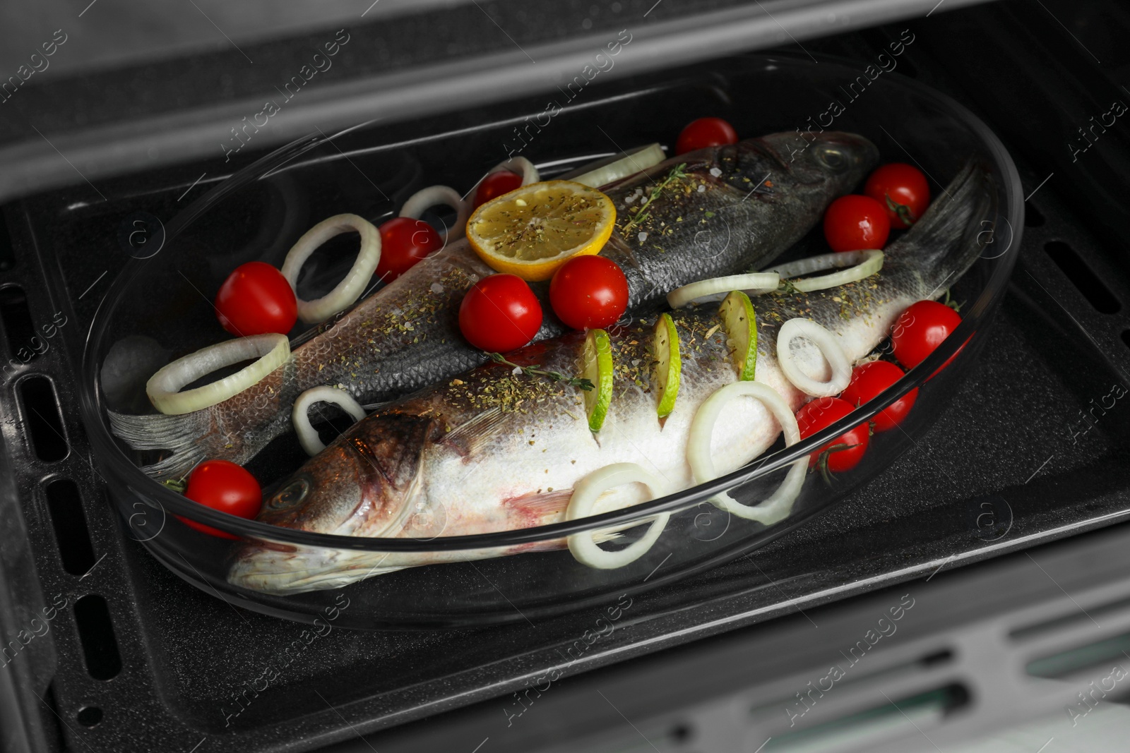 Photo of Glass baking tray with raw sea bass fish and ingredients in oven, closeup