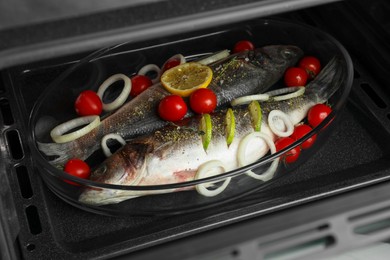 Glass baking tray with raw sea bass fish and ingredients in oven, closeup