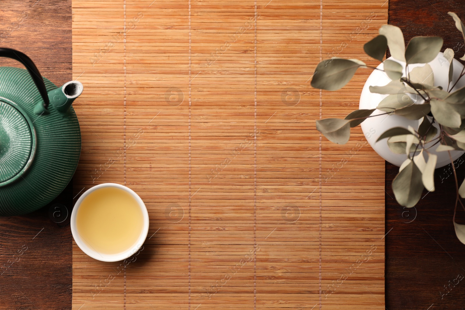 Photo of Bamboo mat, teapot, cup of tea and vase with plant on wooden table, flat lay. Space for text