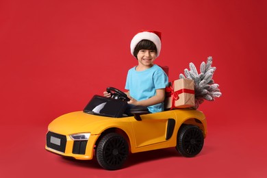 Photo of Cute little boy in Santa hat with Christmas tree and gift box driving children's electric toy car on red background