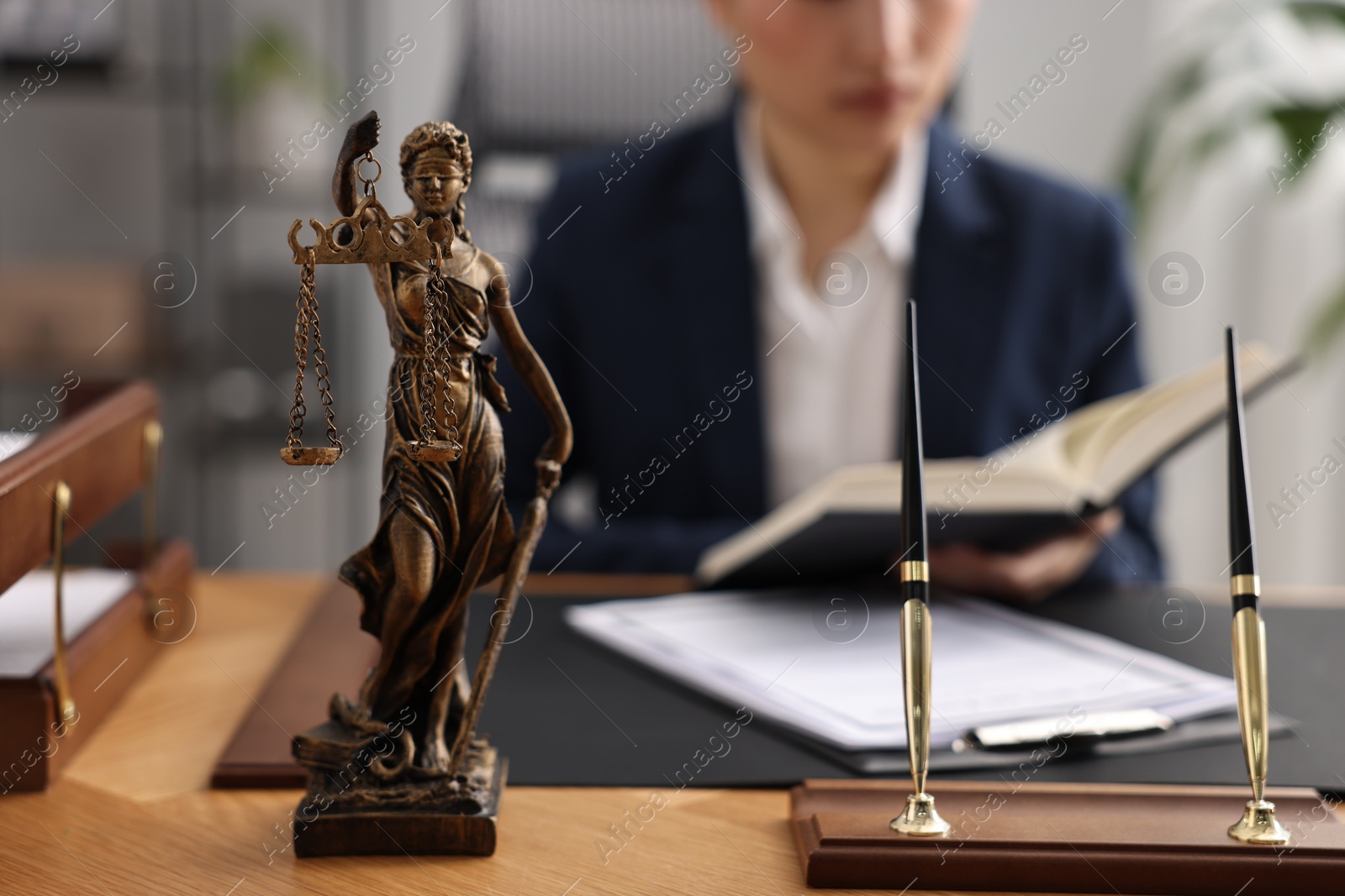 Photo of Notary reading book at table in office, focus on statue of Lady Justice and pens