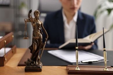 Photo of Notary reading book at table in office, focus on statue of Lady Justice and pens