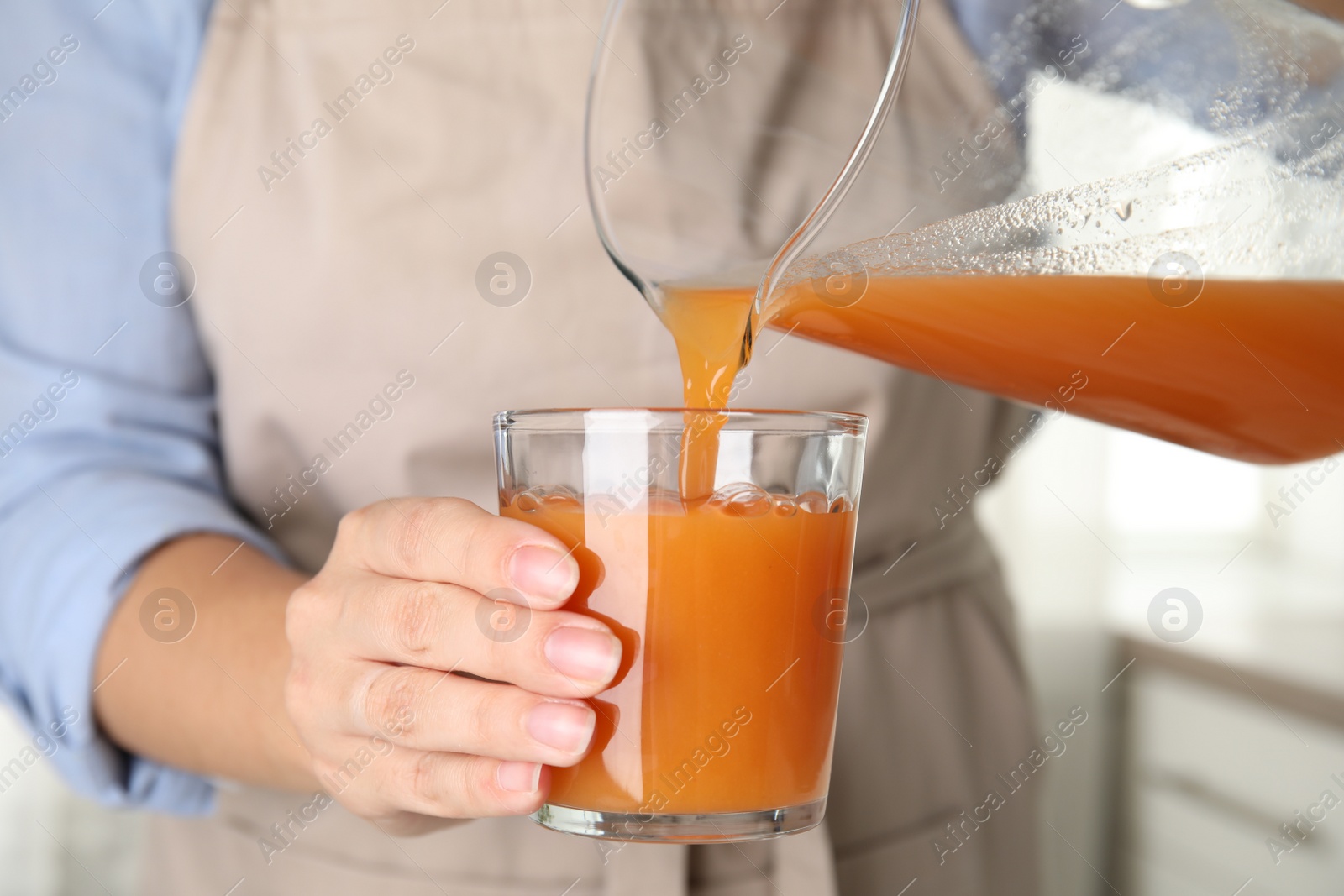 Photo of Woman pouring freshly made carrot juice into glass in kitchen, closeup