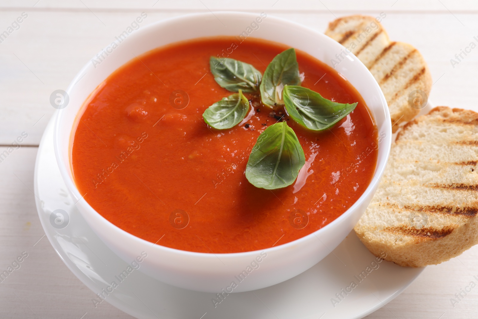 Photo of Delicious tomato cream soup in bowl with pieces of grilled bread on white wooden table, closeup