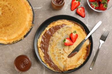 Photo of Delicious thin pancakes served with chocolate paste and strawberries on grey marble table, flat lay