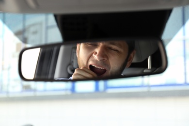 Photo of Tired man yawning in his modern car, through rear view mirror