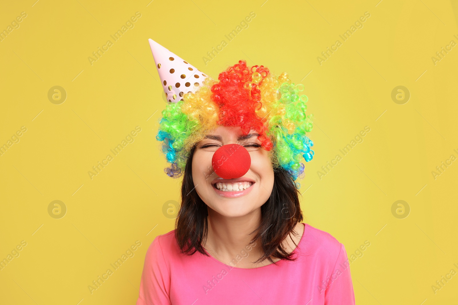 Photo of Joyful woman with rainbow wig, party hat and clown nose on yellow background. April fool's day