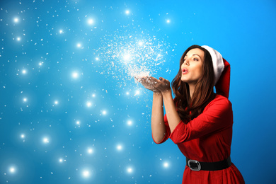 Image of Beautiful woman in Santa hat blowing snow on light blue background, space for text. Christmas party
