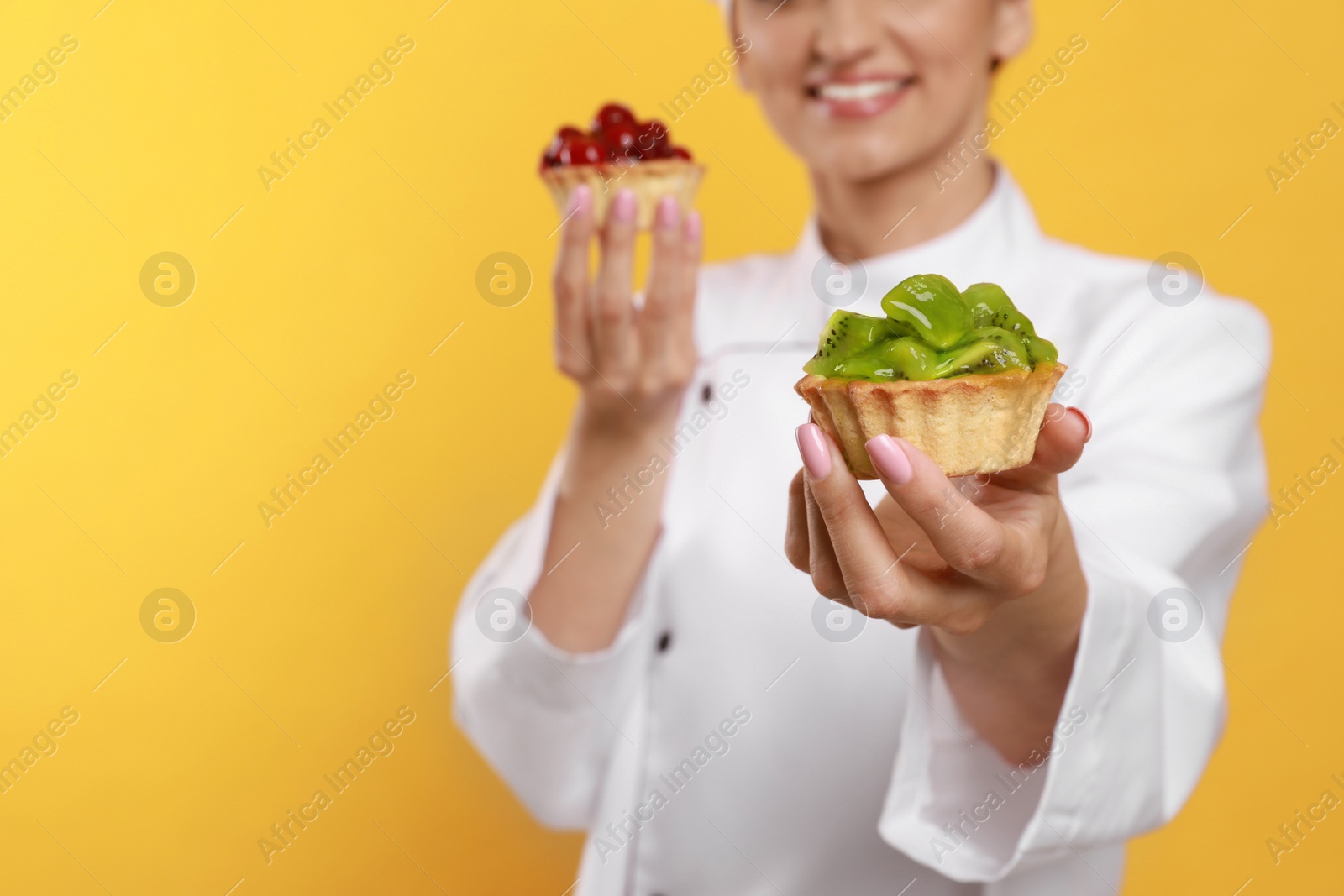 Photo of Happy professional confectioner in uniform with delicious tartlets on yellow background, closeup. Space for text