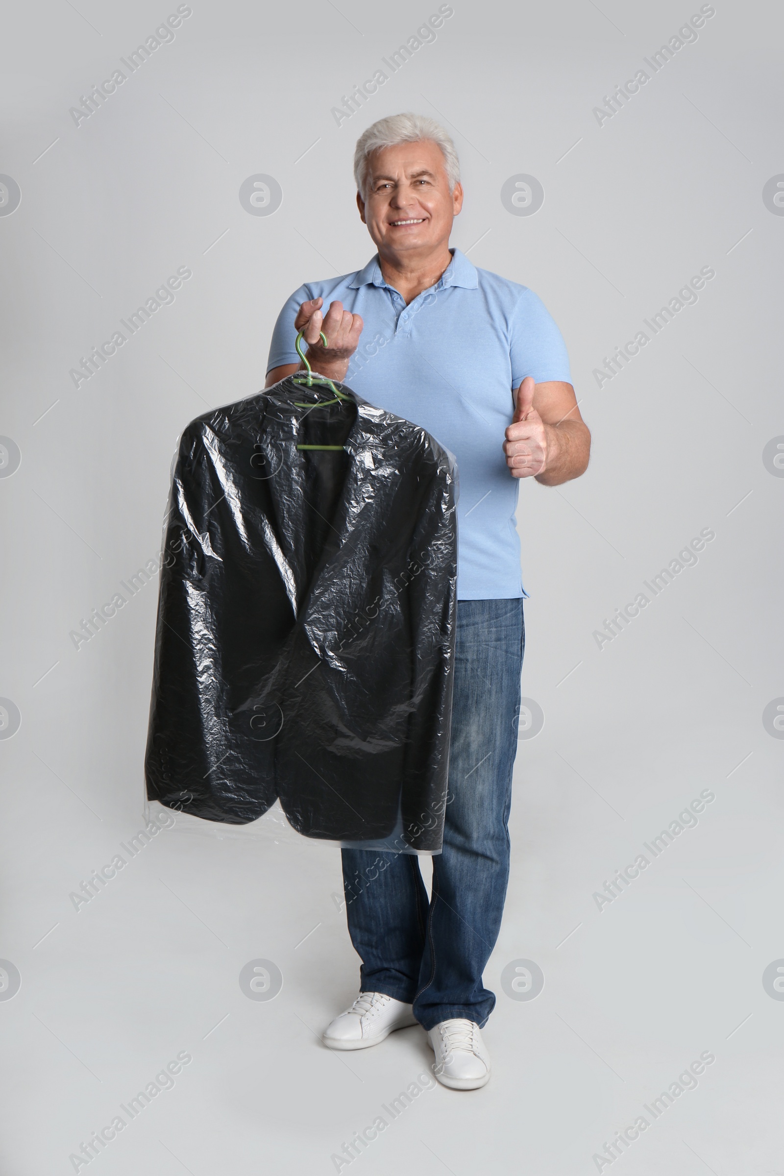 Photo of Senior man holding hanger with jacket in plastic bag on light grey background. Dry-cleaning service