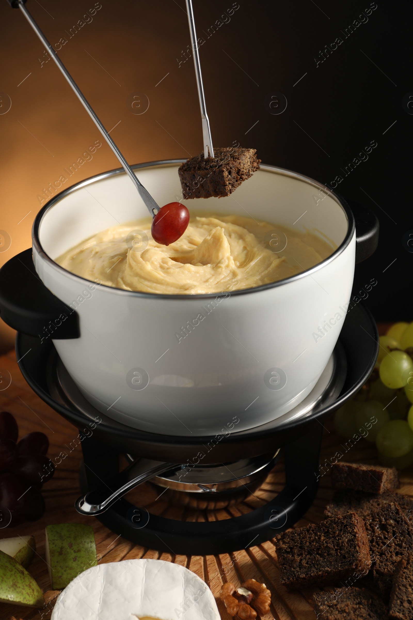 Photo of Dipping pieces of bread and grape into fondue pot with melted cheese at table with snacks, closeup