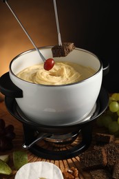 Photo of Dipping pieces of bread and grape into fondue pot with melted cheese at table with snacks, closeup