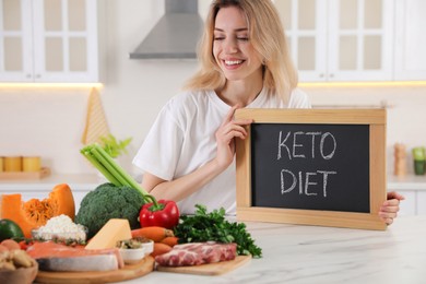 Woman holding blackboard with phrase Keto Diet and different products at white table in kitchen