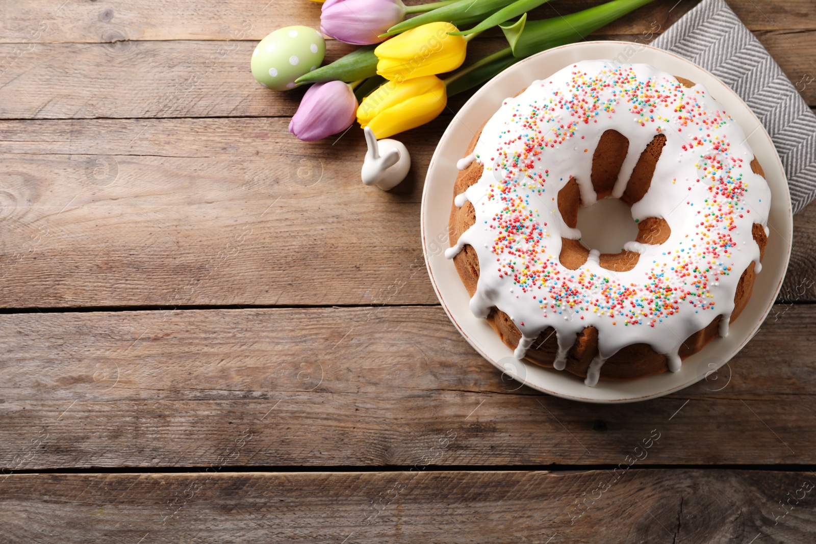 Photo of Glazed Easter cake with sprinkles, decorative bunny, painted egg and tulips on wooden table, flat lay. Space for text