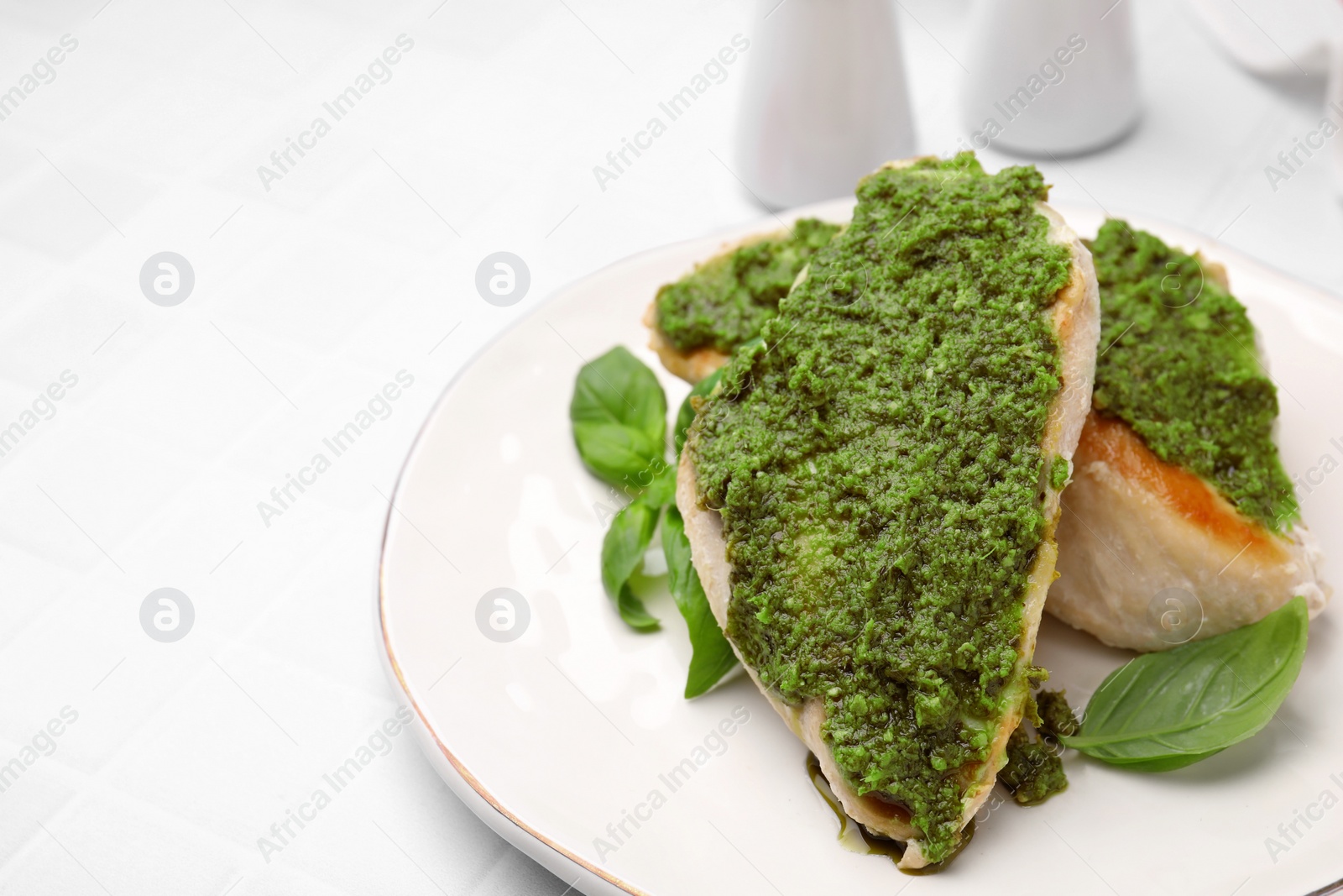 Photo of Delicious chicken breasts with pesto sauce and basil on white tiled table, closeup. Space for text
